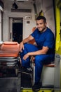 Smiling man in medical uniform sits in the ambulance car beside stretcher