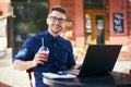 Smiling man with plastic cup of fresh juice in hand works with laptop. Businessman in glasses drinks beverage for body