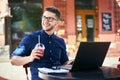 Smiling man with plastic cup of fresh juice in hand works with laptop. Businessman in glasses drinks beverage for body