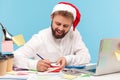 Smiling man in santa claus hat writing down letter or signing greeting postcard in red envelope, romance way to congratulate with