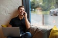 Smiling man using his laptop and phoning on sofa at window Royalty Free Stock Photo