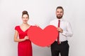 Smiling man in white shirt, woman in red dress standing together, holding big heart, expressing love Royalty Free Stock Photo