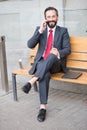 Smiling manager sitting on bench and phoning. Handsome smiled young businessman siting on bench with his laptop next to office