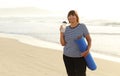 Smiling mature adult woman with bottle of water after exercising outdoors holding yoga mat outdoors on beach by sea on Royalty Free Stock Photo