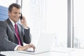 Smiling mature businessman talking on cell phone while using laptop in office Royalty Free Stock Photo