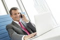 Smiling mature businessman talking on cell phone while using laptop in office Royalty Free Stock Photo