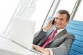 Smiling mature businessman talking on cell phone while using laptop in office Royalty Free Stock Photo