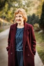 Smiling mature caucasian woman walking in autumn park outdoors CIS in light burgundy coat