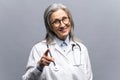 Smiling mature gray-haired female doctor in white medical gown isolated on grey Royalty Free Stock Photo