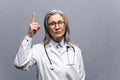 Smiling mature gray-haired female doctor in white medical gown, with stethoscope isolated on grey background points Royalty Free Stock Photo