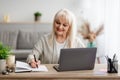 Smiling mature lady writing in notebook using laptop at home