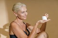 Smiling mature woman in black bra smiling and looking at white jar of cream beauty product in her hands, posing half Royalty Free Stock Photo