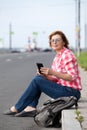 Smiling mature woman sitting on sloping curb, smartphone in her hands, looking far away, empty road Royalty Free Stock Photo