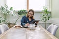Smiling mature woman working at home, having breakfast and using digital tablet, business notebook Royalty Free Stock Photo