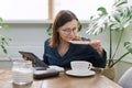 Smiling mature woman working at home, having breakfast and using digital tablet, business notebook Royalty Free Stock Photo