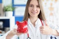 Smiling medical doctor woman holding red pig piggy bank in her hands and gesture thumbs up Royalty Free Stock Photo