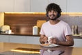 A smiling mestizo guy stands in the kitchen in subdued lighting, holds a cup of tea or coffee, listens to music on