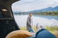 Smiling middle-aged man dressed in warm knitted clothes and jeans near the car. Trunk mountain lake view with woman legs in white