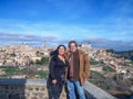 Smiling middle-aged tourist couple standing at viewpoint against blue sky, Toledo cityscape in background