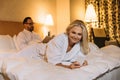 smiling middle aged woman in bathrobe lying on bed and using smartphone while husband reading Royalty Free Stock Photo