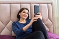 Smiling middle-aged woman in headphones looking into a smartphone, lying in bed at home Royalty Free Stock Photo