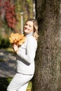 Smiling middle-aged woman with long wavy fair hair in roll-neck sweater holding bouquet of fallen leaves leaning on tree Royalty Free Stock Photo