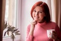 Smiling middle-aged woman with red hair standing with white mug near window. Short coffee break concept Royalty Free Stock Photo