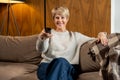 Smiling middle-aged woman sitting on the sofa and turning on the TV with a remote control Royalty Free Stock Photo