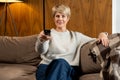 Smiling middle-aged woman sitting on the sofa and turning on the TV with a remote control Royalty Free Stock Photo
