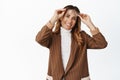 Smiling middle aged woman take on eyewear and looking happy, trying new glasses, standing in brown suit against white Royalty Free Stock Photo