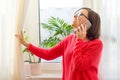 A smiling middle-aged woman is talking on the phone, opening the curtains and looking out at the sunny window. Side view. Royalty Free Stock Photo