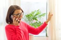 Smiling middle-aged woman is talking on the phone, opening the curtains and looking out at the sunny window. Royalty Free Stock Photo