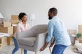 Smiling millennial african american male and female carry armchair in room with cardboard boxes with things