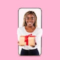 Smiling millennial black female gives box with gift from empty screen of huge phone, isolated on pink background