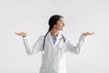 Smiling millennial indian woman doctor in white coat raises her hands and makes choice, isolated on light background