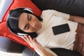 Smiling mixed race gender fluid man lying on couch wearing wireless headphones using smartphone Royalty Free Stock Photo