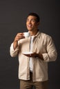 Mixed race man in beige shirt looking away and holding cup of coffee and saucer on black background