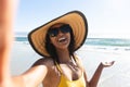 Smiling mixed race woman wearing sunhat and sunglasses taking selfie on beach Royalty Free Stock Photo