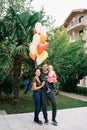 Smiling mom hugging dad with a little girl in his arms in the garden near colorful balloons Royalty Free Stock Photo