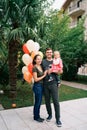 Smiling mom hugging dad with a little girl in his arms while standing in the garden near balloons Royalty Free Stock Photo