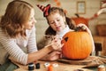 Smiling mother and child girl drawing scary faces on Halloween pumpkins while sitting in cozy kitchen