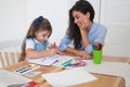 Smiling mother and daughter preparing for lessons and draws at the table with pencils and paints. Parent and pupil of preschool.