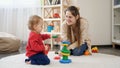 Smiling mother playing with her baby son on carpet and giving him colroful toys Royalty Free Stock Photo