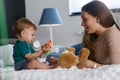 Smiling mother playing with her cute toddler son at home in the bed. Using plush bear and baby toys. Maternity concept Royalty Free Stock Photo