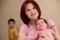 Smiling mom posing with infant child. Older kid is standing left behind, out of focus. Parenting issues when new baby arrives