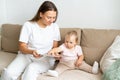 Smiling mother reading book with curious baby girl on sofa in living room. Horizontal natural light Royalty Free Stock Photo