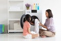 Smiling mother sitting looking at her cute daughter helping her loading clothes into a washing machine in a laundry room at home. Royalty Free Stock Photo