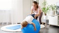 Smiling mother in sports uniform stretching and training her baby son on fitball at home. Concept of child healthcare, kids sports Royalty Free Stock Photo