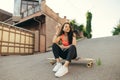 Smiling mulatto girl sitting on logboard on street background, looking down, wearing street clothes. Girl skater relaxing on Royalty Free Stock Photo