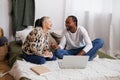 Smiling multiethnic couple talking while using laptop near book on bed, online education concept Royalty Free Stock Photo